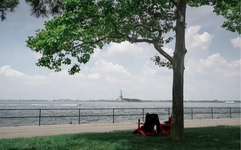Things to Do in NYC 11 Red chairs under a tree on Governors Island with the Statue of Liberty across the harbor