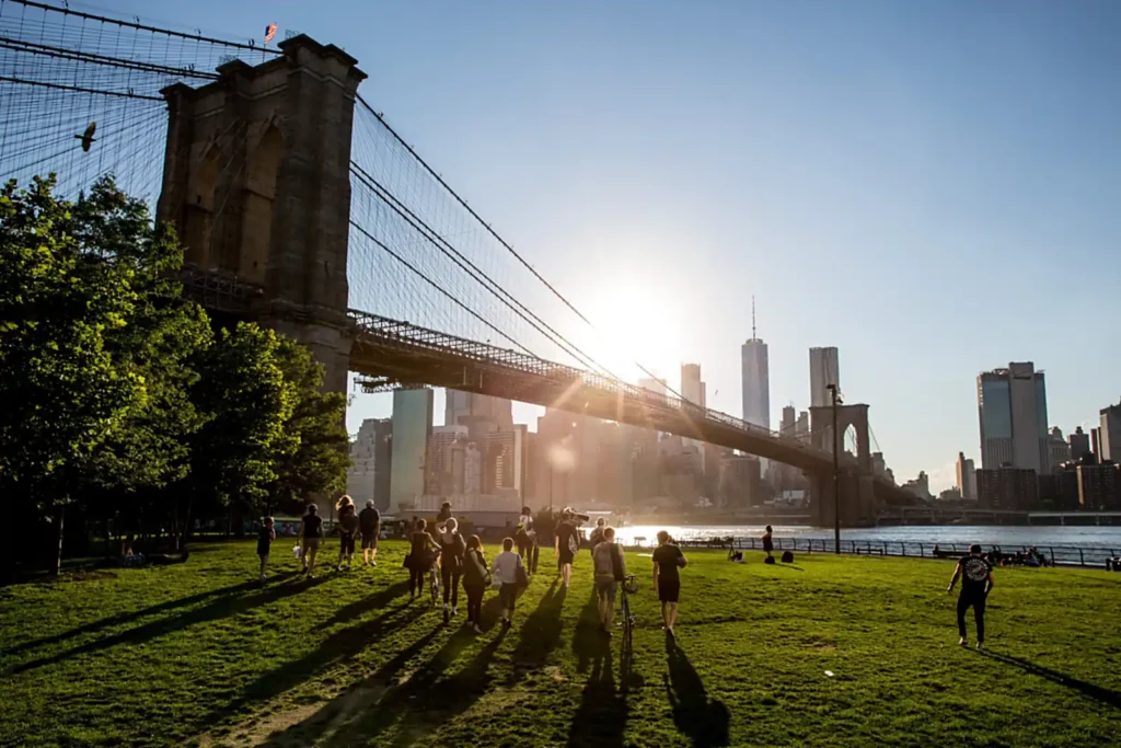 Brooklyn Bridge Park: Every Pier, What's Free, and When Things Are Actually Open 4 People walking across a lawn in Brooklyn Bridge Park at sunset, with the Brooklyn Bridge and Lower Manhattan behind