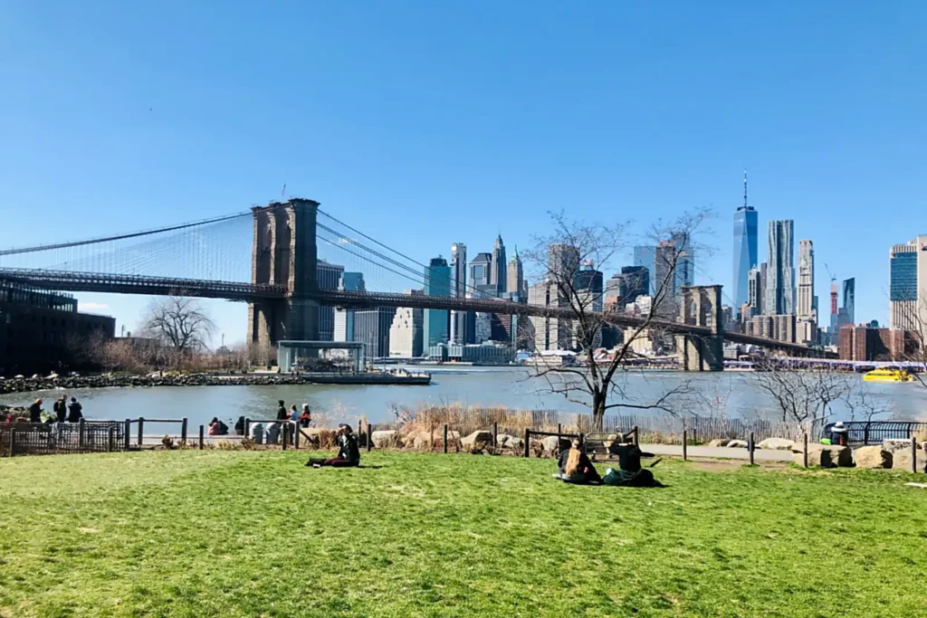 Brooklyn Bridge Park: Every Pier, What's Free, and When Things Are Actually Open 2 People sitting on a lawn in Brooklyn Bridge Park with the Brooklyn Bridge and Lower Manhattan skyline behind