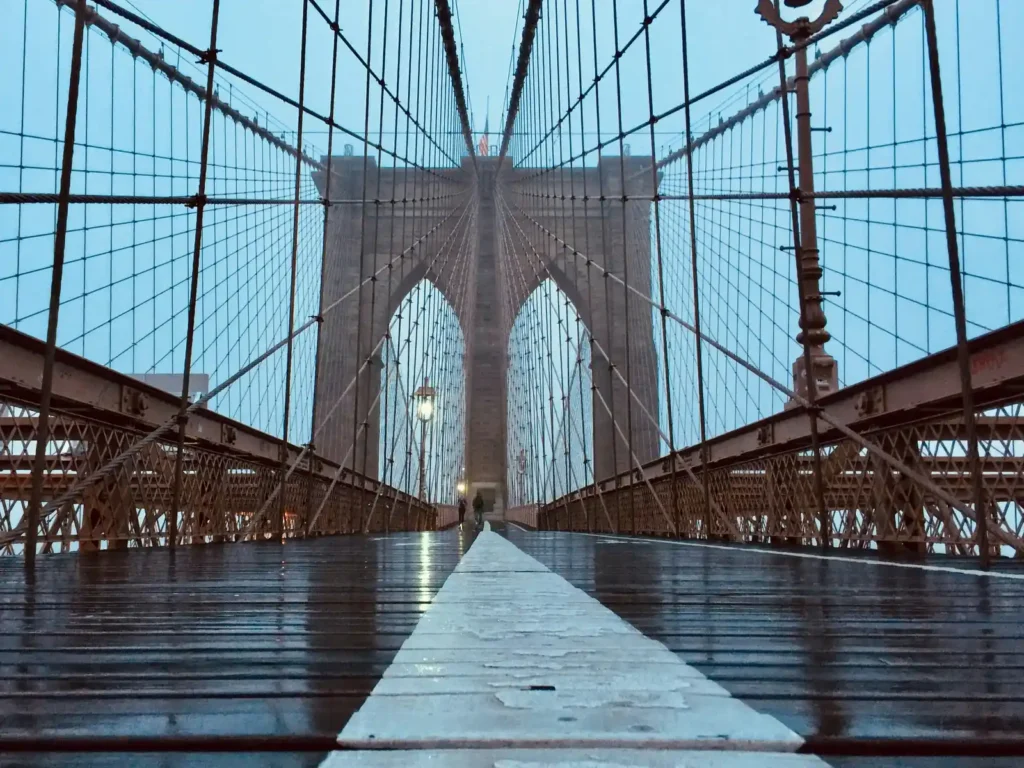 Walking the Brooklyn Bridge: Entrances, Timing, and What Changed in 2026 2 The wooden pedestrian promenade on the Brooklyn Bridge, looking toward the stone tower at dawn