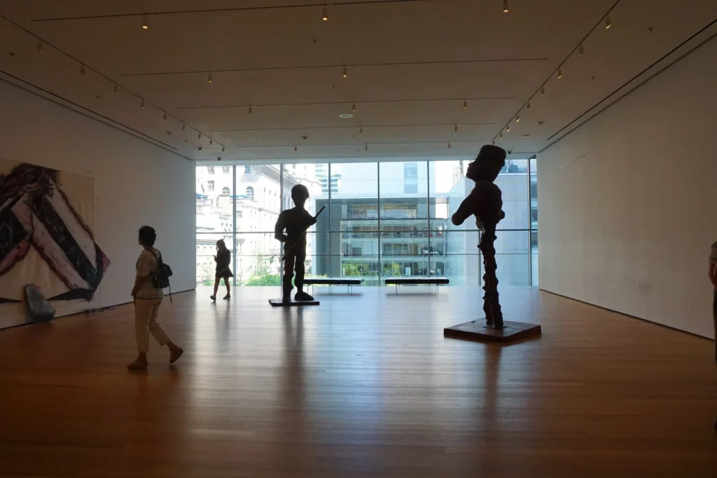 MoMA: Tickets, Free Friday, and Every Other Way In 2 Visitors walking through a MoMA collection gallery, with sculptures silhouetted against a window onto Midtown