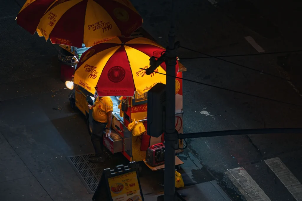 Cheap Eats in NYC: What Street Food Actually Costs 1 A Halal Guys cart lit up on a Manhattan street corner, its umbrellas reading serving the street since 1990