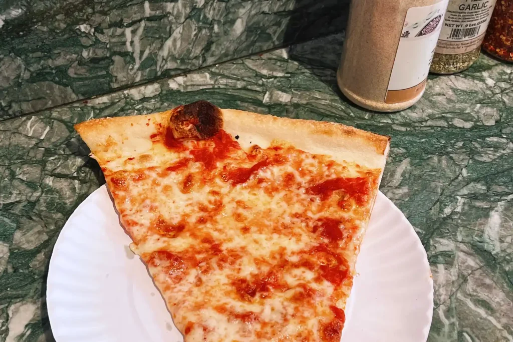 Cheap Eats in NYC: What Street Food Actually Costs 2 A plain cheese slice on a paper plate at a New York pizza counter, with garlic and oregano shakers behind