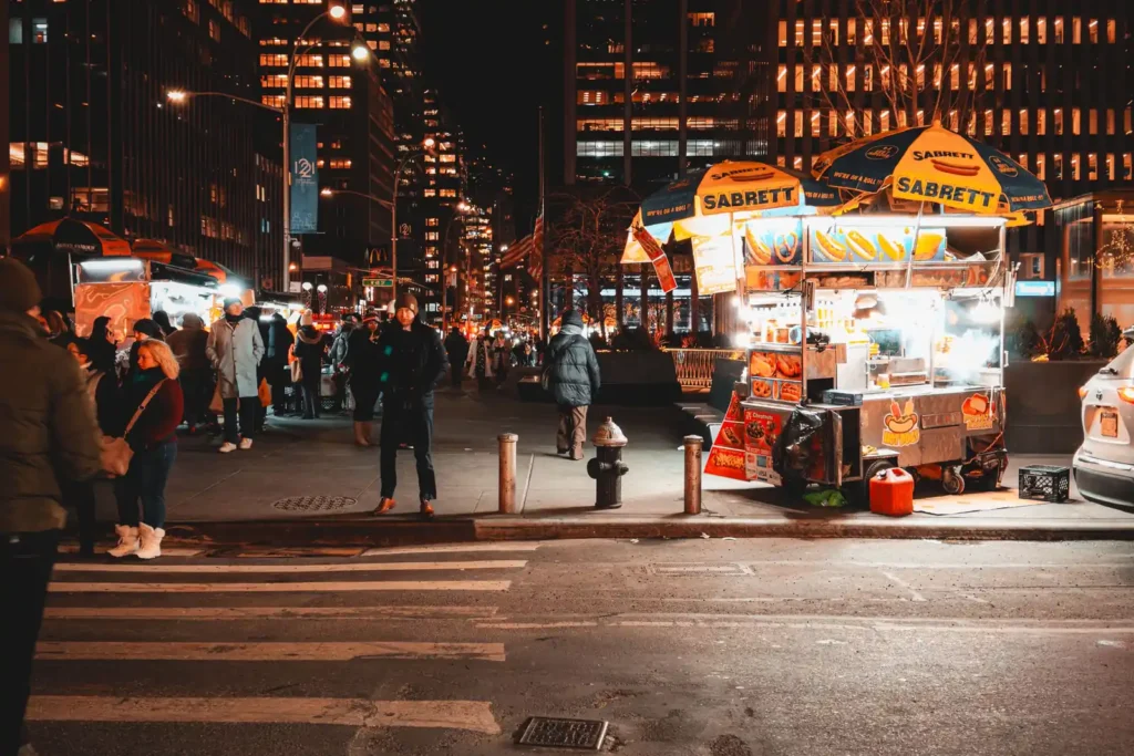 Cheap Eats in NYC: What Street Food Actually Costs 4 A lit hot dog cart on a Manhattan street corner at night, with other food carts and office towers behind