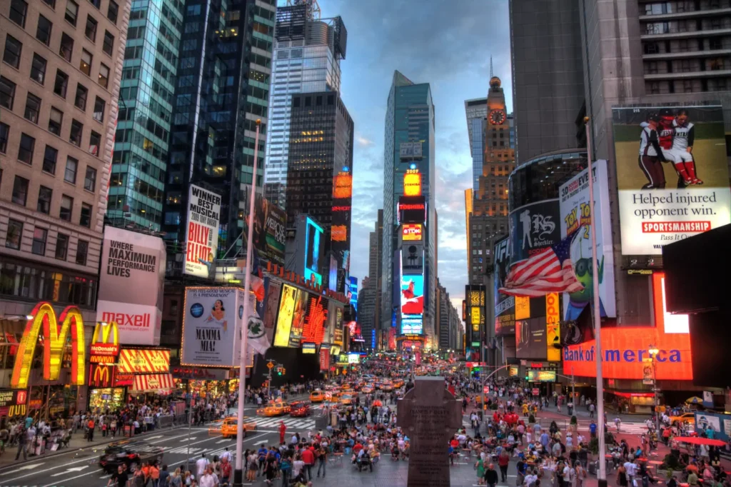 Times Square: Where It Is, What Is Worth Seeing, and How to Do It at Night 4 Times Square at dusk looking south from the northern end toward One Times Square, with billboards lit against a blue evening sky and crowds on the plaza
