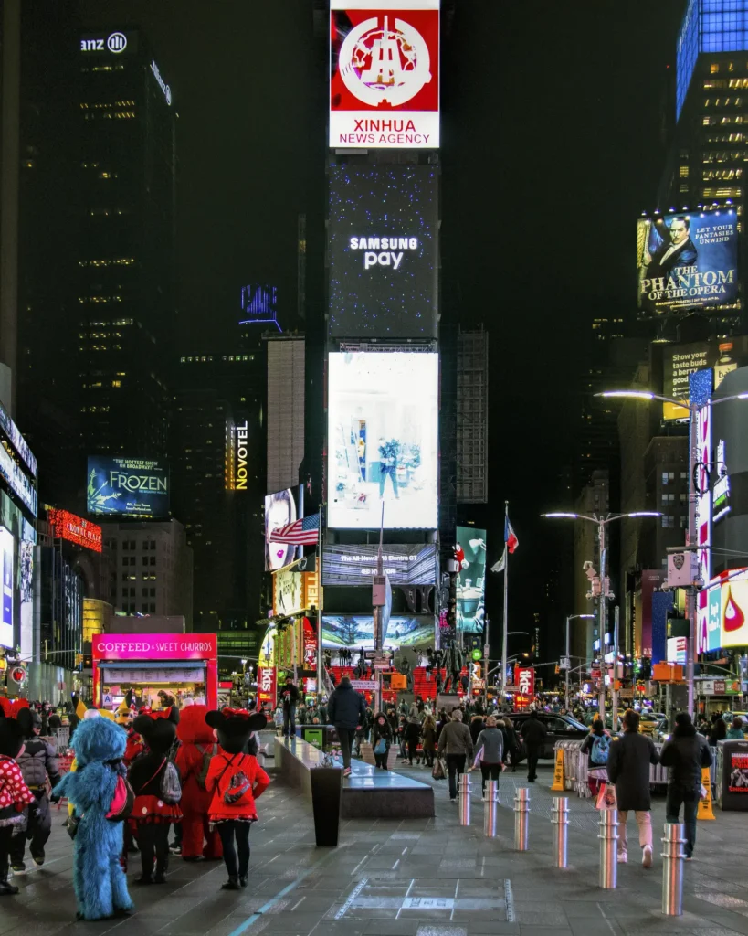 Times Square: Where It Is, What Is Worth Seeing, and How to Do It at Night 3 Costumed characters on the Times Square plaza at night, with the Red Steps, the TKTS booth and a tower of billboards in the distance